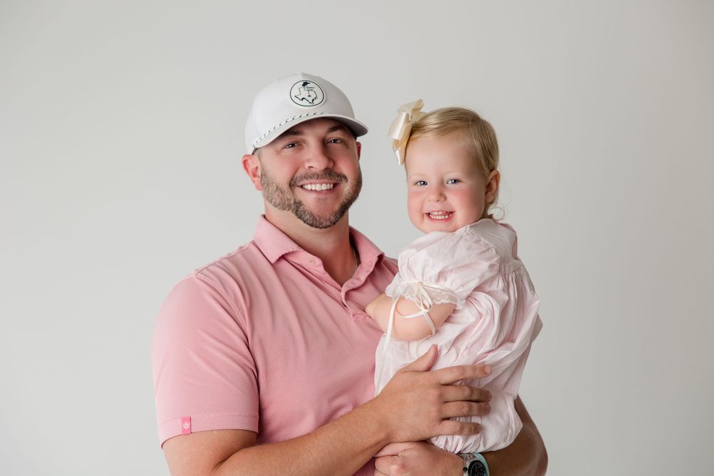 A child in a white dress smiles with family members wearing pink clothing during a photo session.