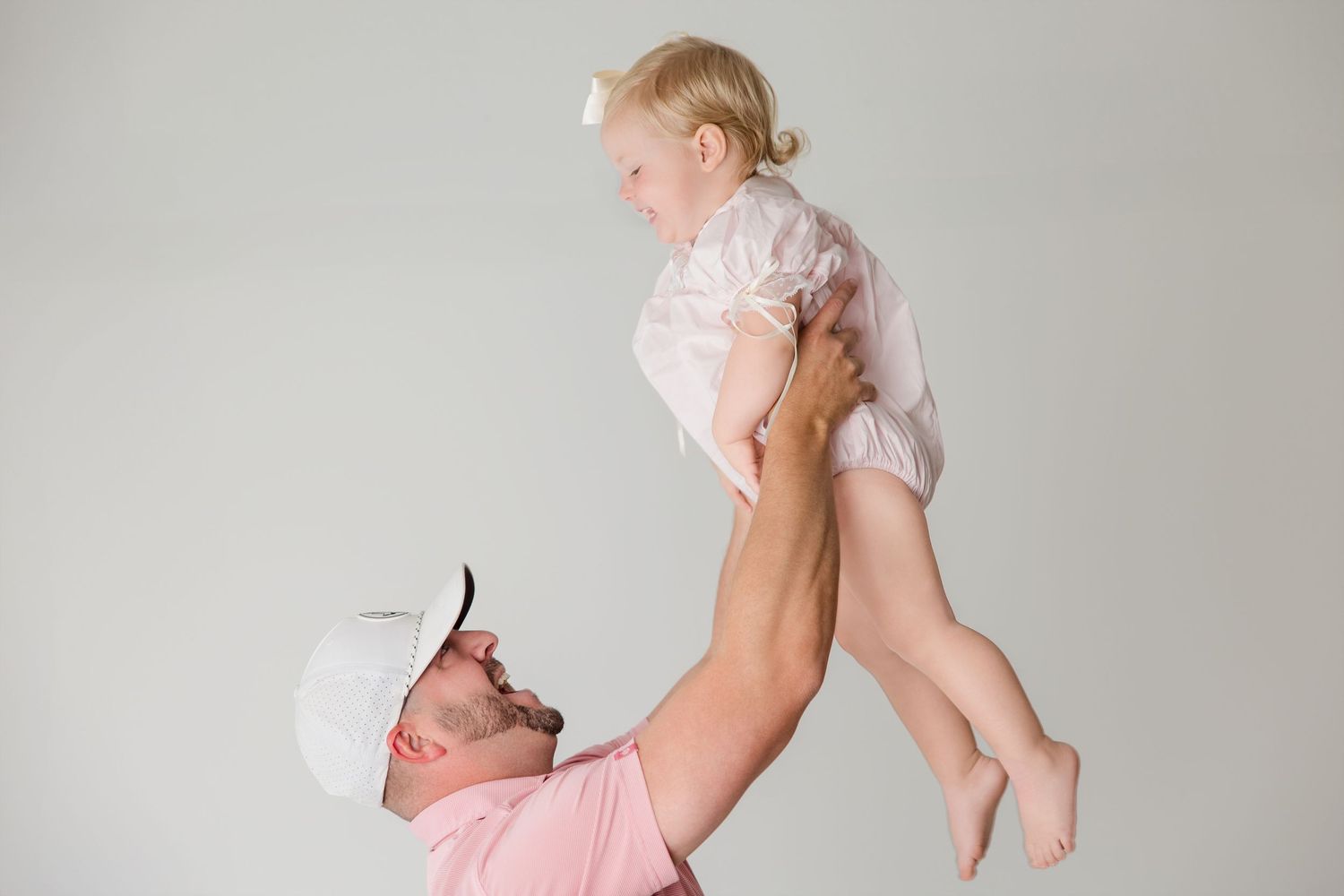 A person wearing pink holds up a happy baby in a white onesie against a light gray background.