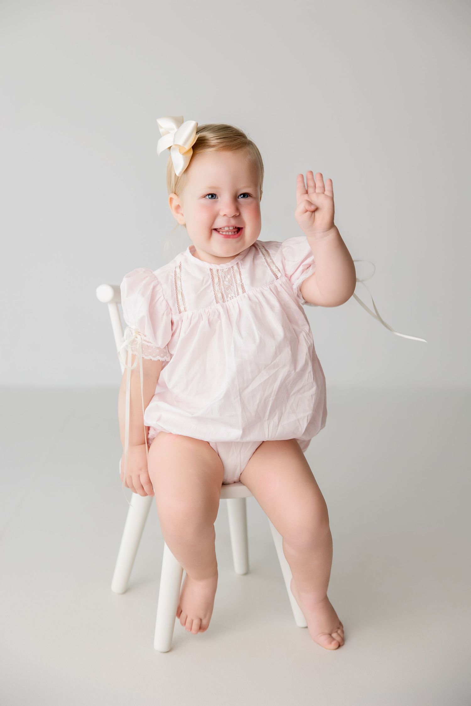 Laughing baby wearing a pink dress while sitting in a white chair poses for a portrait series.