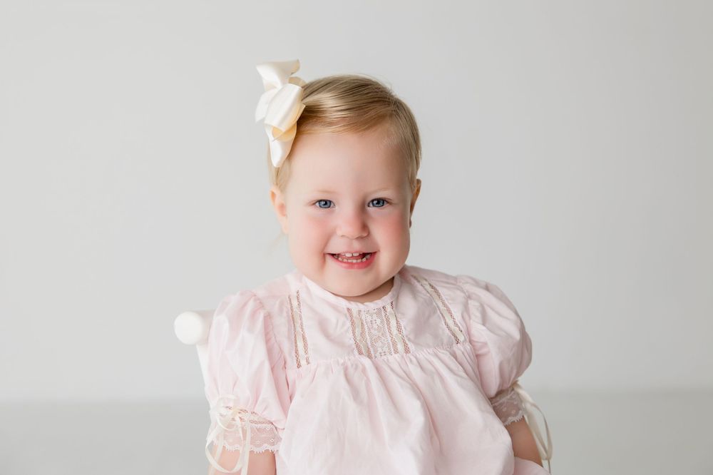 A child in a white lace dress with a bow smiles sweetly during a portrait session.