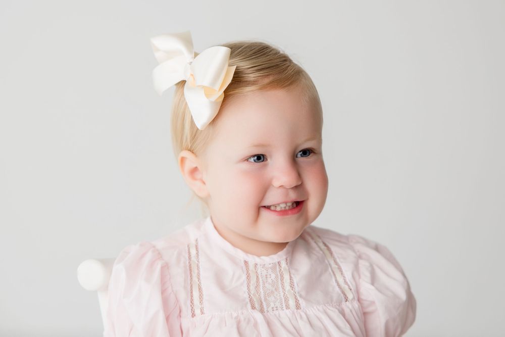 A beautiful baby wearing a light pink dress with a white bow hair accessory smiles delightfully against a white backdrop.