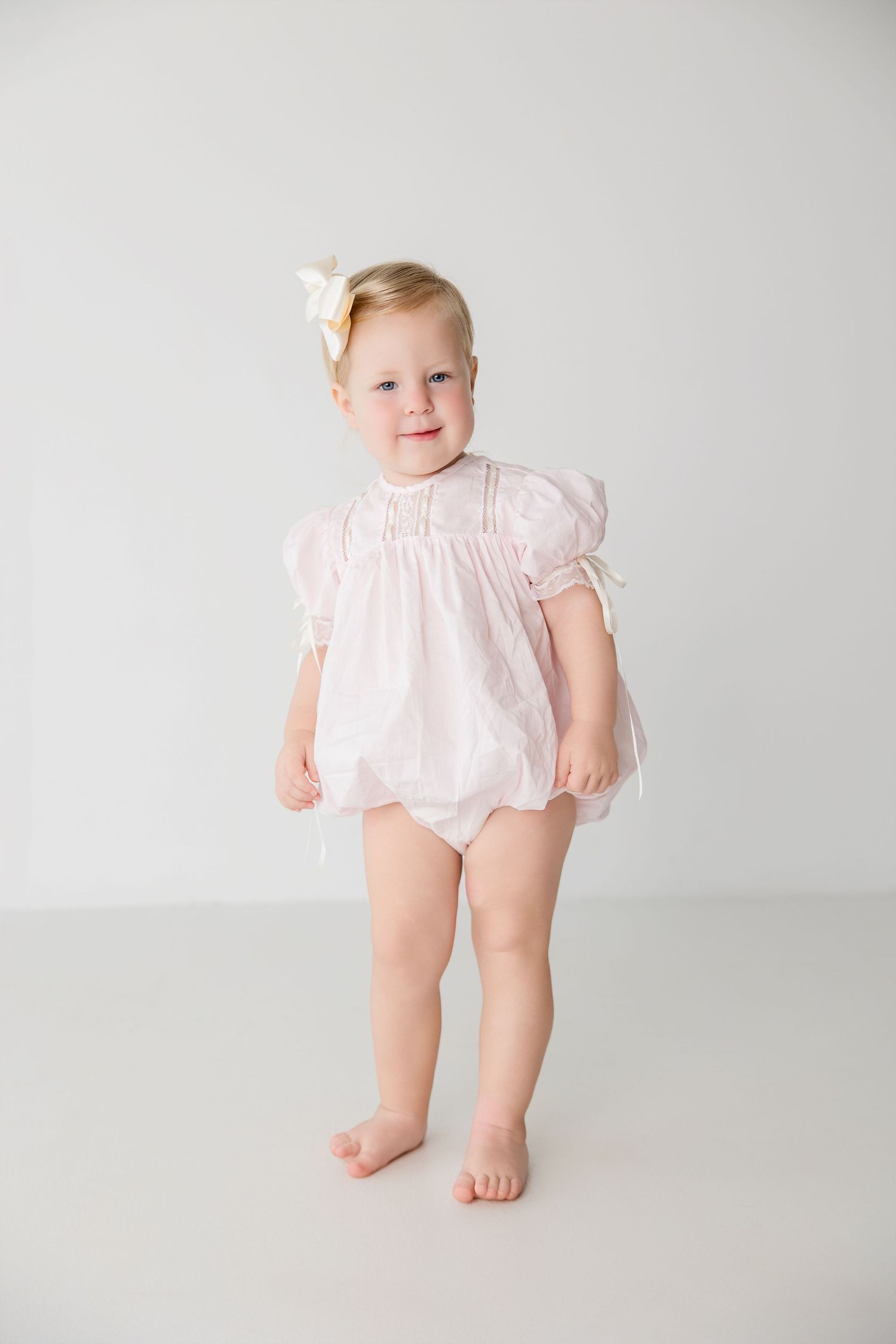 Portrait of a barefoot toddler wearing a light pink smocked dress with puffy sleeves against a white background.