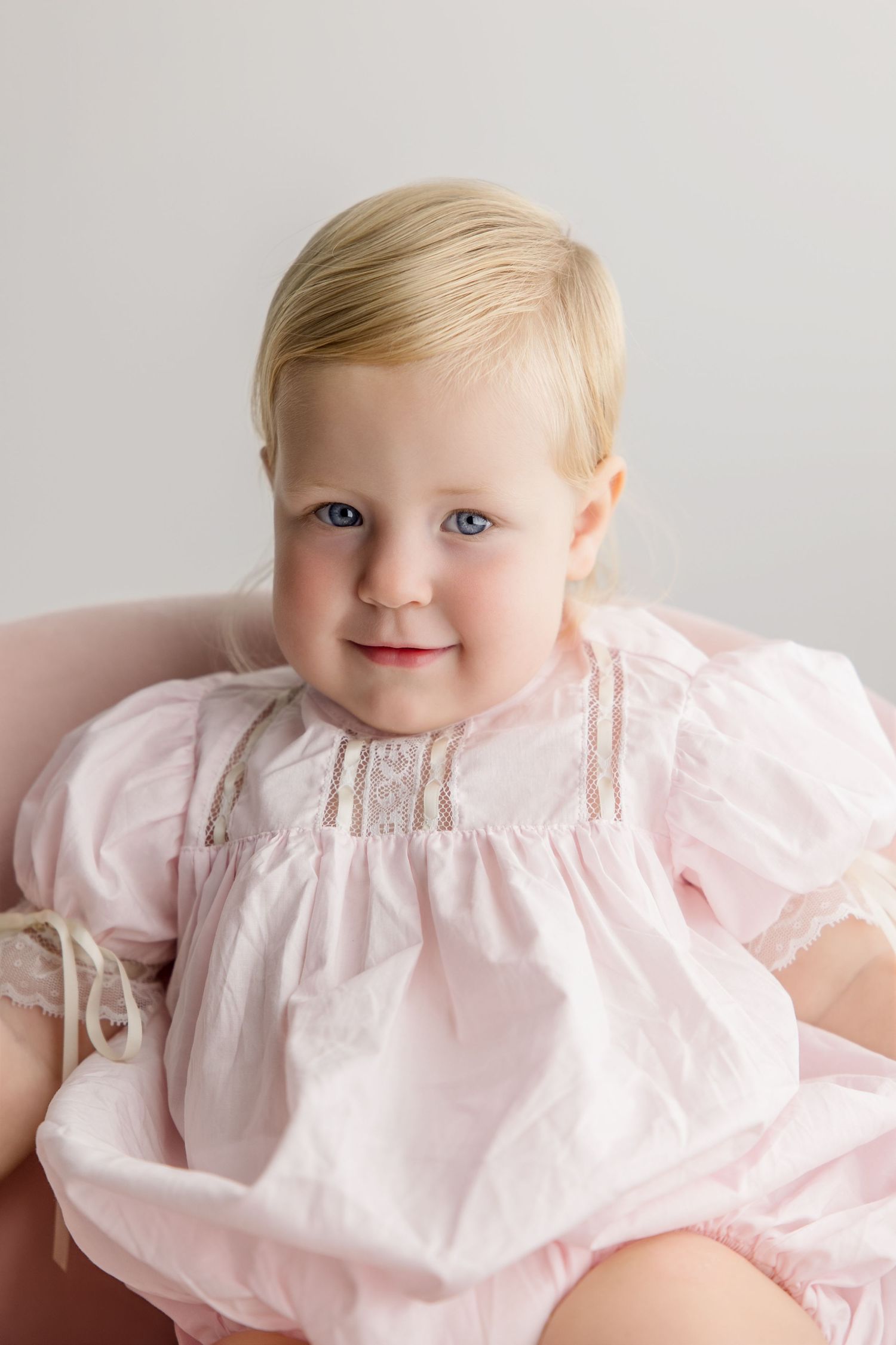 A baby wearing a light pink smock dress with delicate embroidery details sits against a neutral background.
