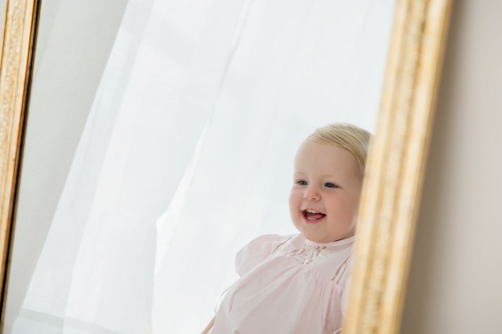 Portrait of a smiling toddler in a pink dress looking towards a window with golden curtains.