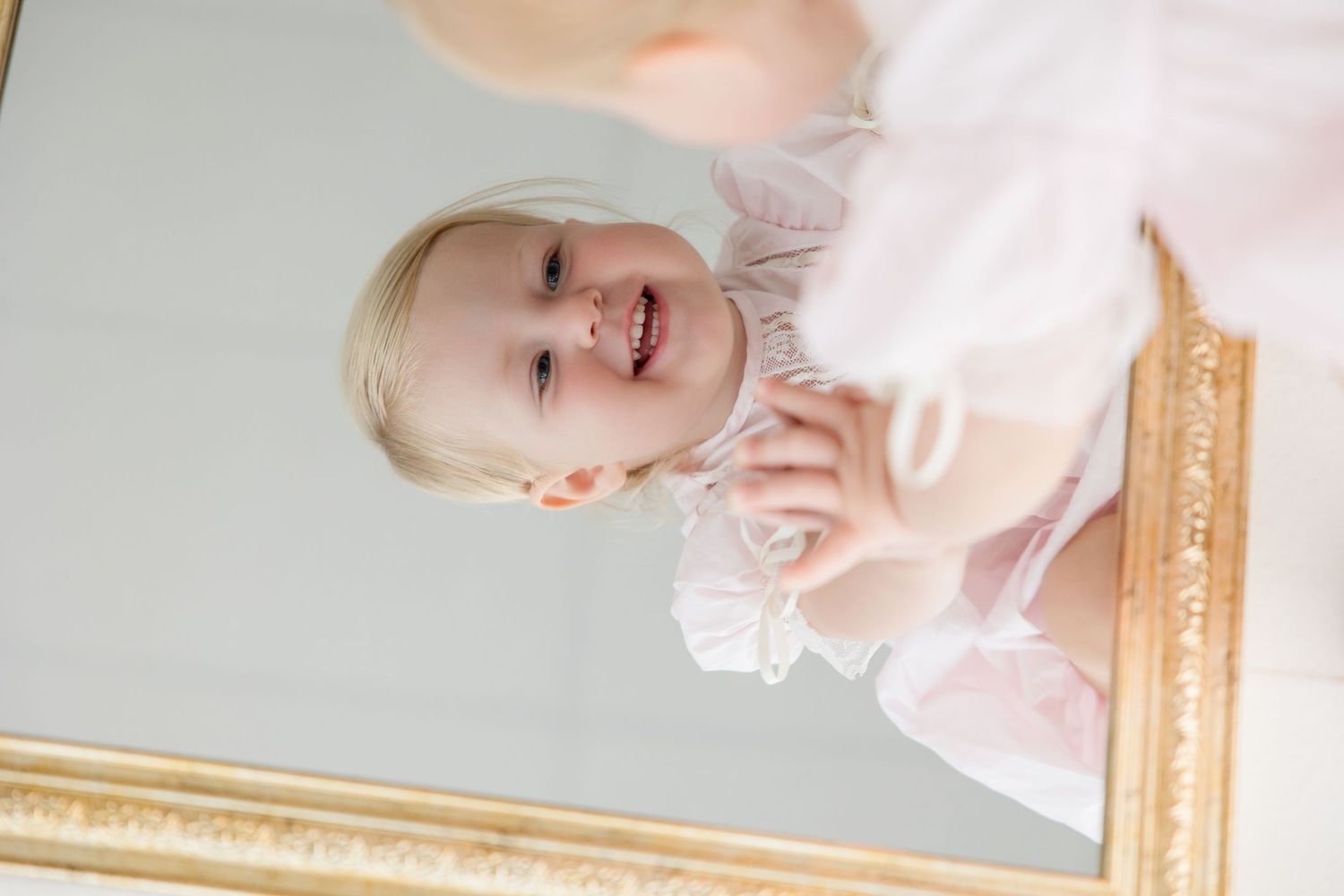 Baby in a pink romper with delicate lace details in front of a mirror creating multiple reflections.