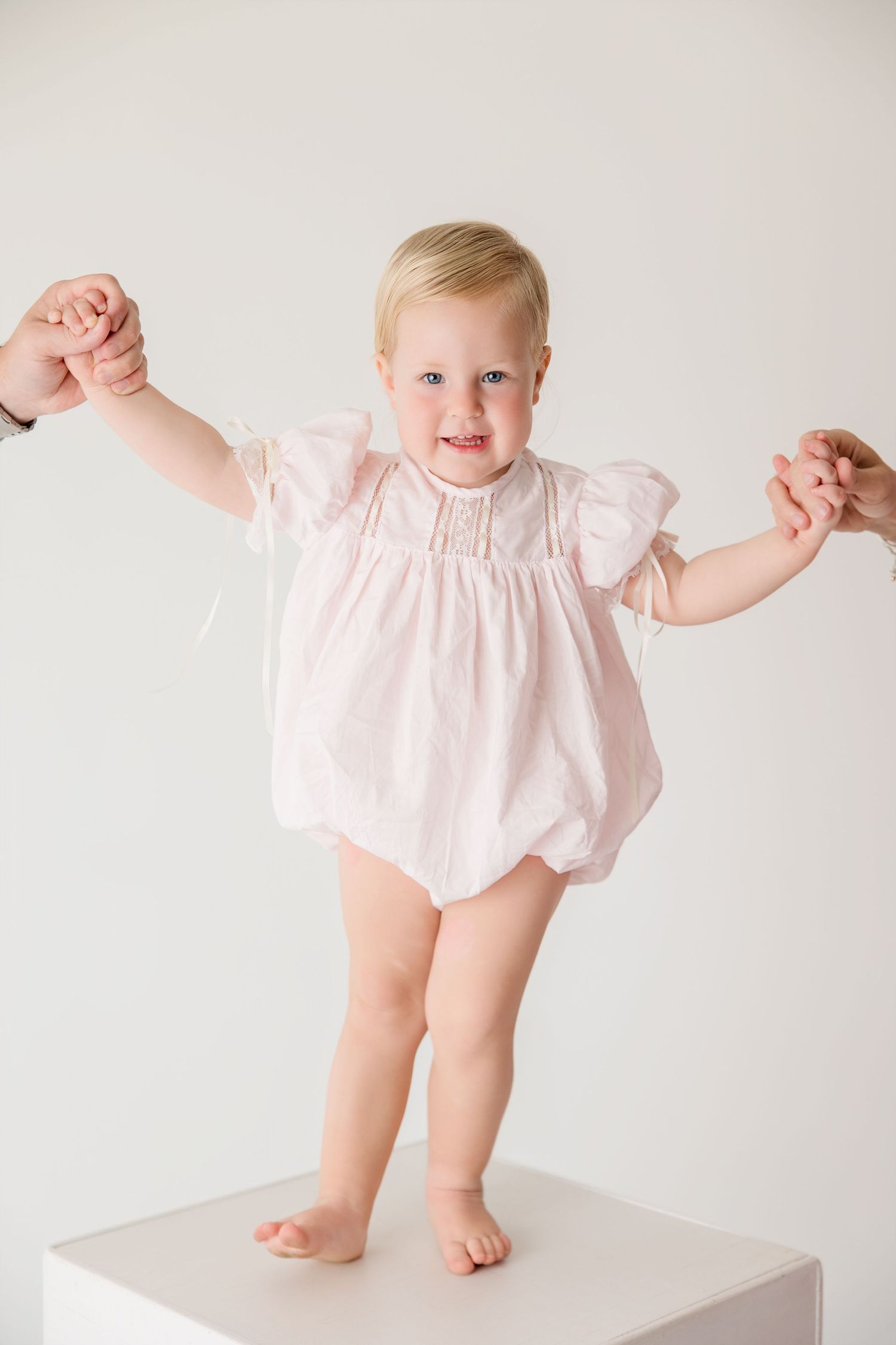 Baby wearing pink romper smiles while standing on white surface in bright studio setting.