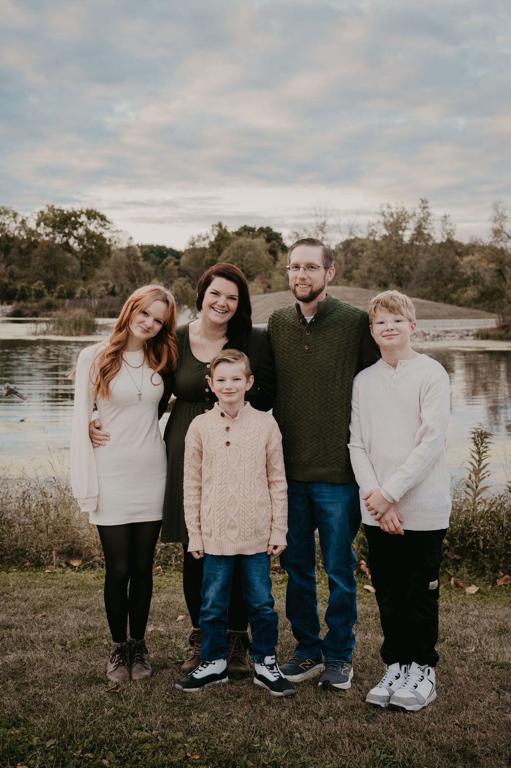 Family of five smiles together near a lake on a cloudy autumn day.