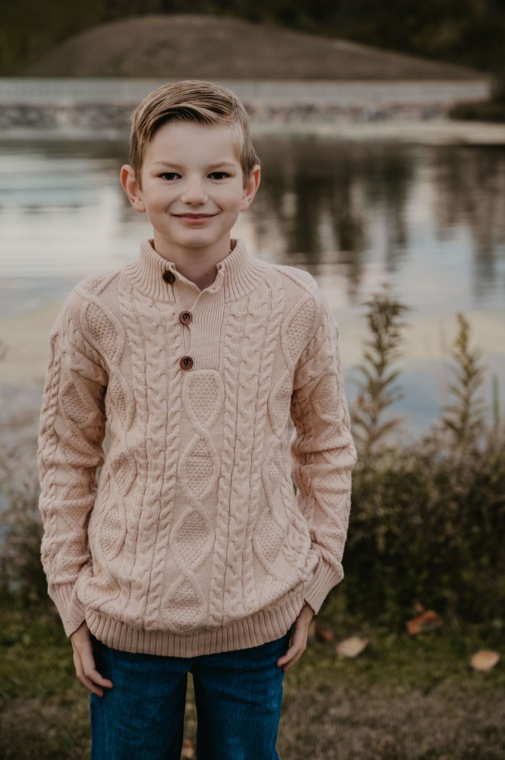 Young boy in beige cable-knit sweater smiles by a lake in autumn.