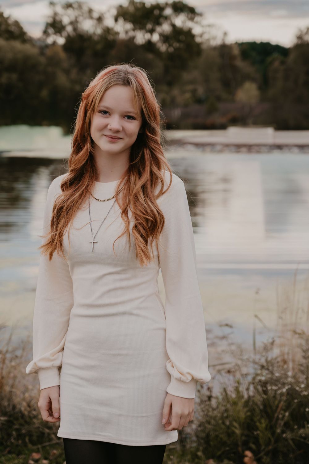Teen girl with long red hair in white dress poses by a lake wearing a cross necklace.