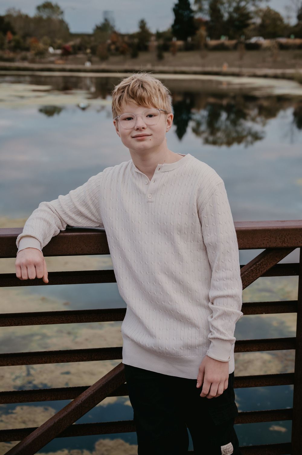 Smiling teenage boy with glasses leaning on a railing with a reflective lake in the background.