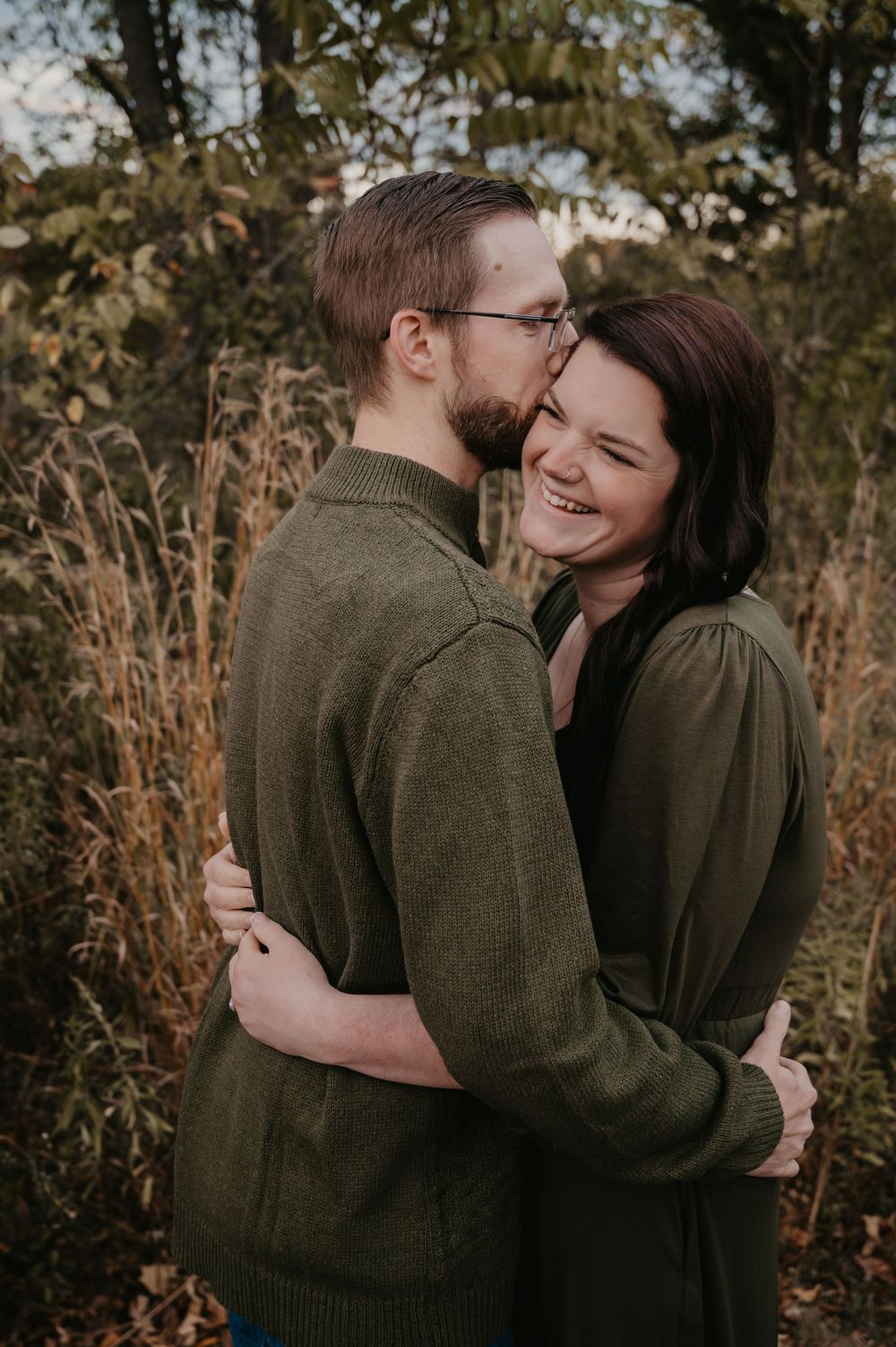 Couple in matching green sweaters embracing and laughing in a field of tall golden grasses.