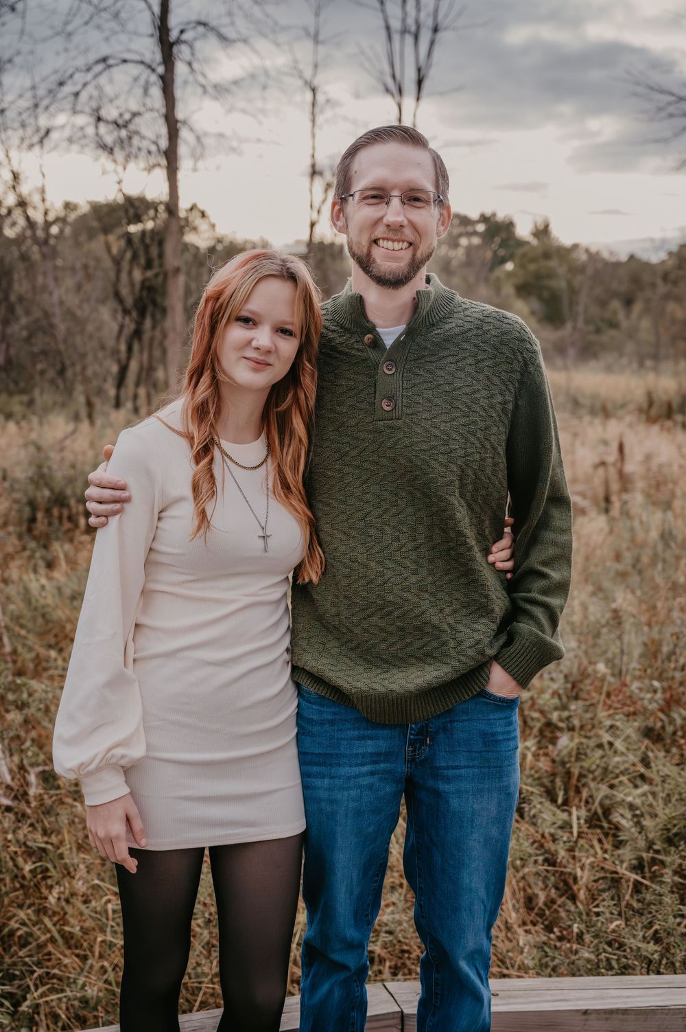 Smiling couple poses outdoors in fall, woman in white dress and man in green sweater and jeans.
