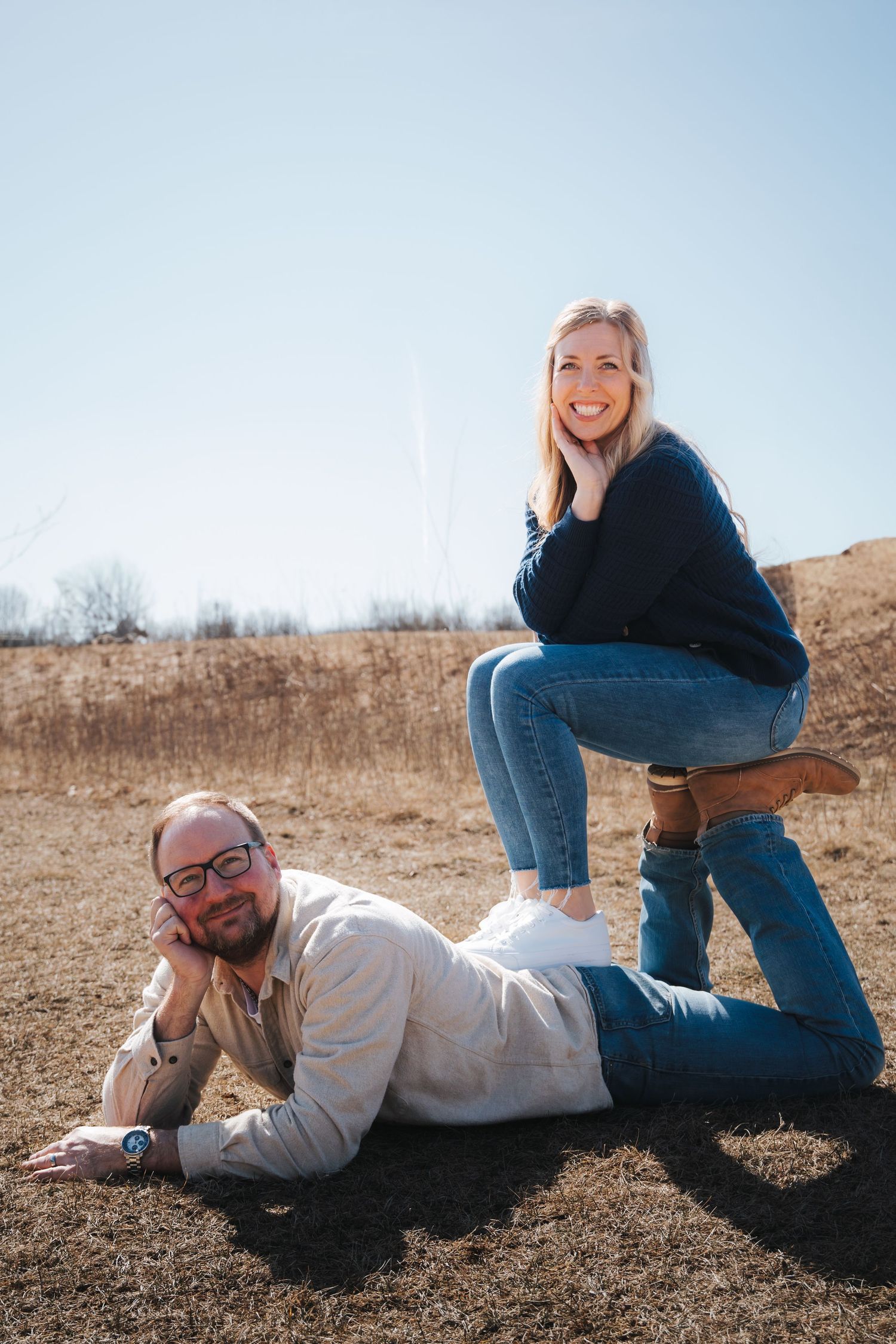 Playful couple lying together on dried grass field during outdoor photoshoot on sunny day.