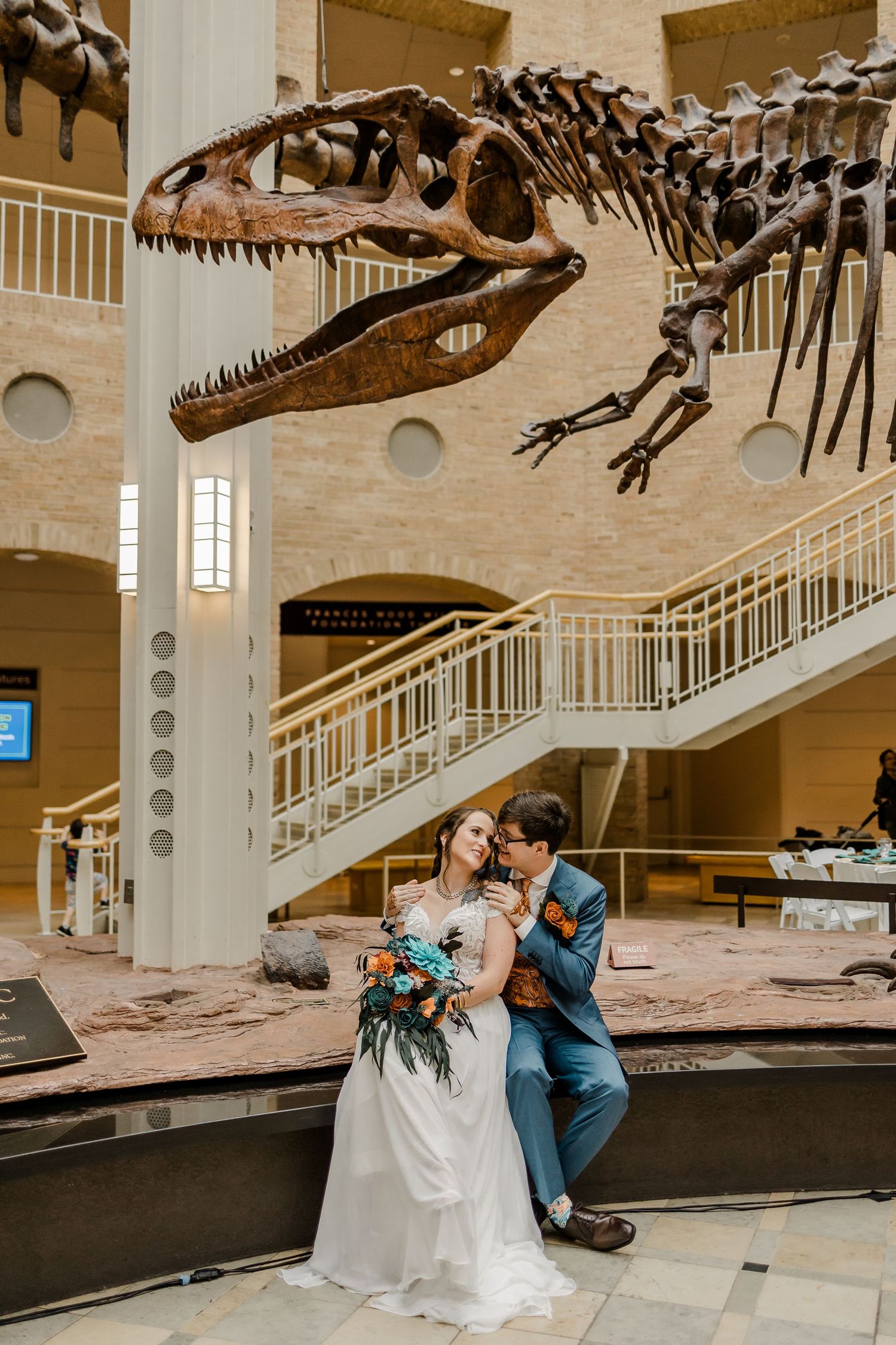 Wedding couple poses with dinosaur skeletons in a natural history museum exhibit hall.