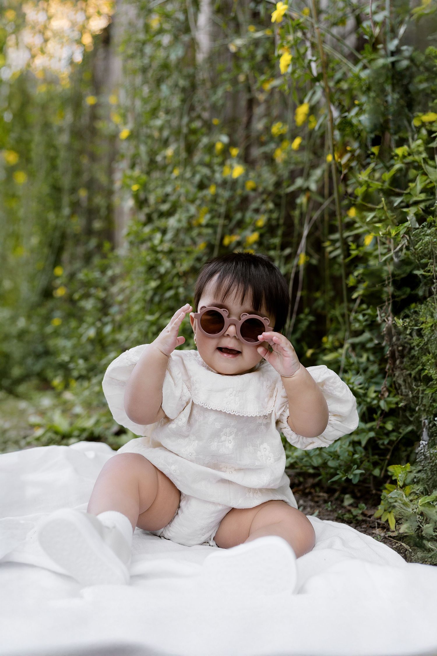 Baby in white dress laughs and plays while sitting on grass with scattered flower petals.