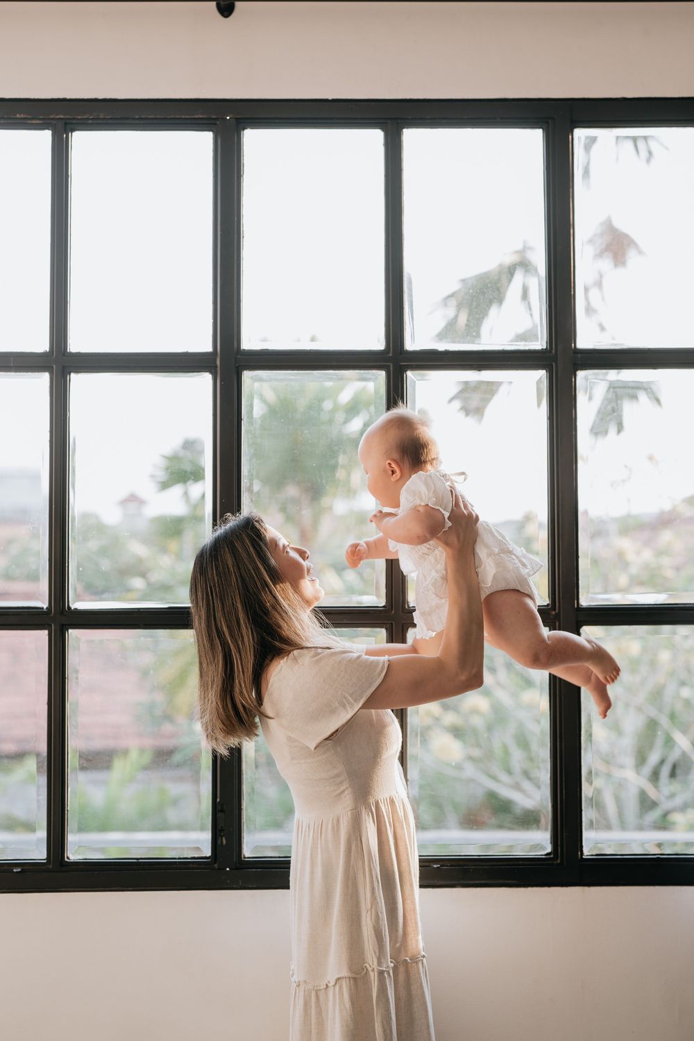 Parent holds baby while standing by black-framed windows overlooking tropical scenery.