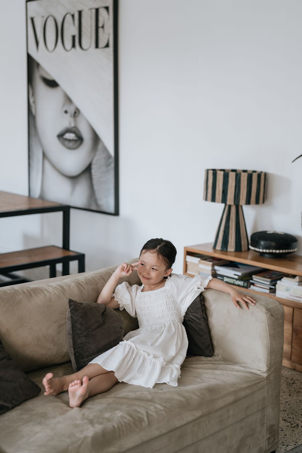 Child in white dress lounging on gray couch with Vogue artwork displayed on wall.