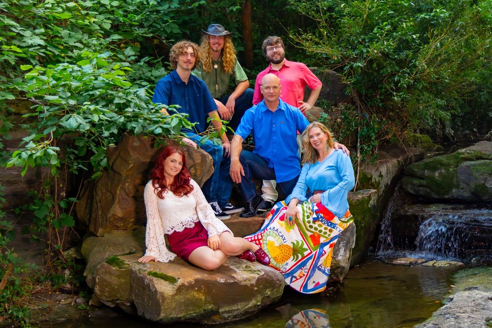 A group of people in colorful attire gather around a small waterfall and rocks in a lush green forest setting.