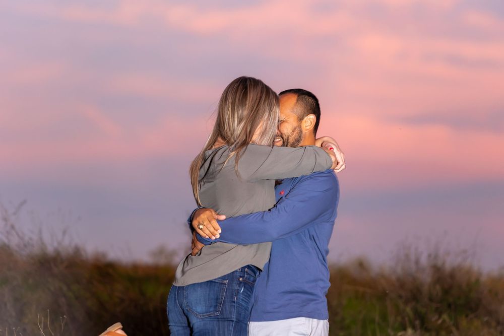 A couple embraces in a warm hug against a beautiful pink sunset sky in a field.
