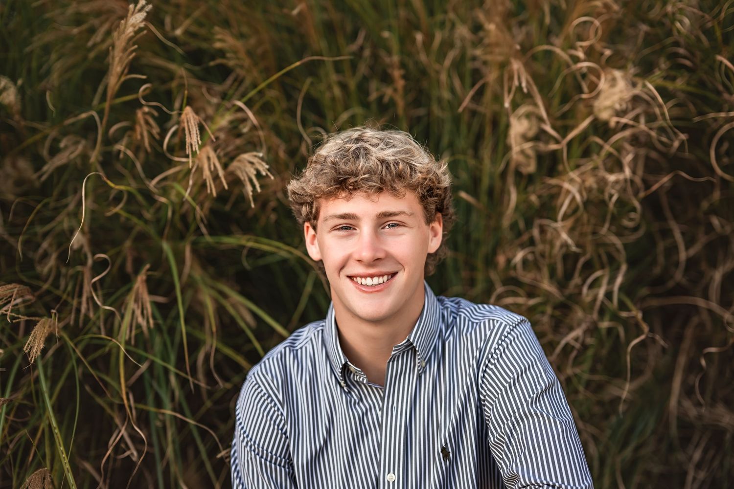smiling headshot of a senior boy in a blue striped shirt surrounded by green and brown foliage.