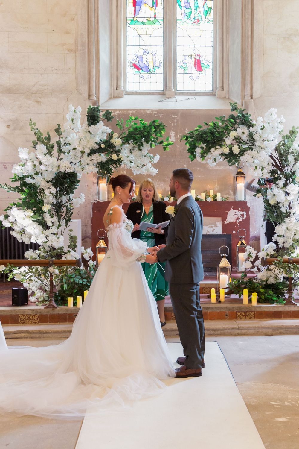 A traditional church wedding ceremony with white floral arrangements and stained glass windows in the background.
