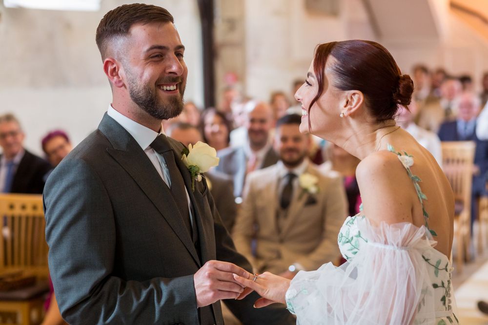 A joyful couple exchanges rings during their wedding ceremony while surrounded by guests in a wooden-pewed venue.