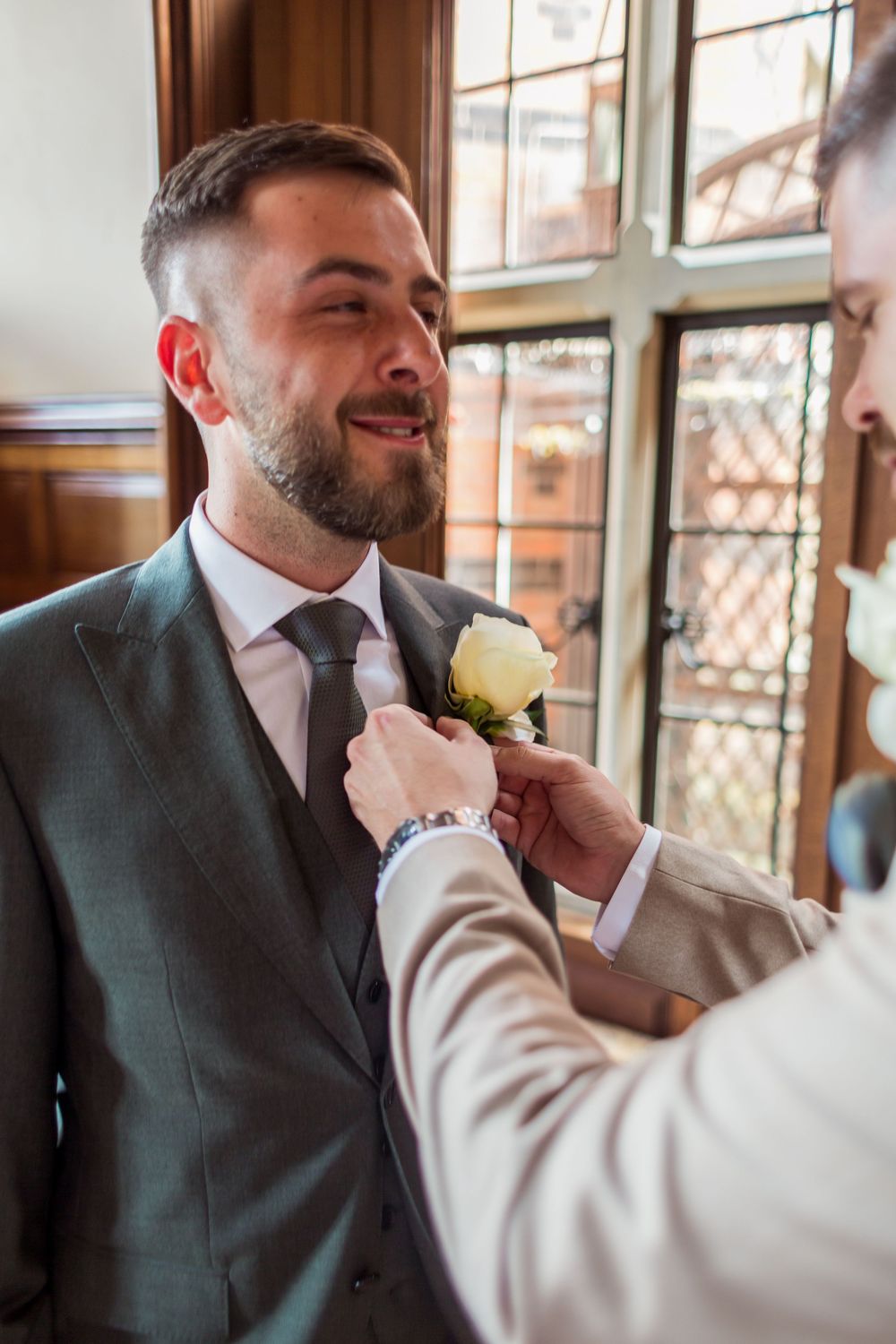 Someone helps pin a white rose boutonniere onto a grey suit jacket during a formal event by a window.