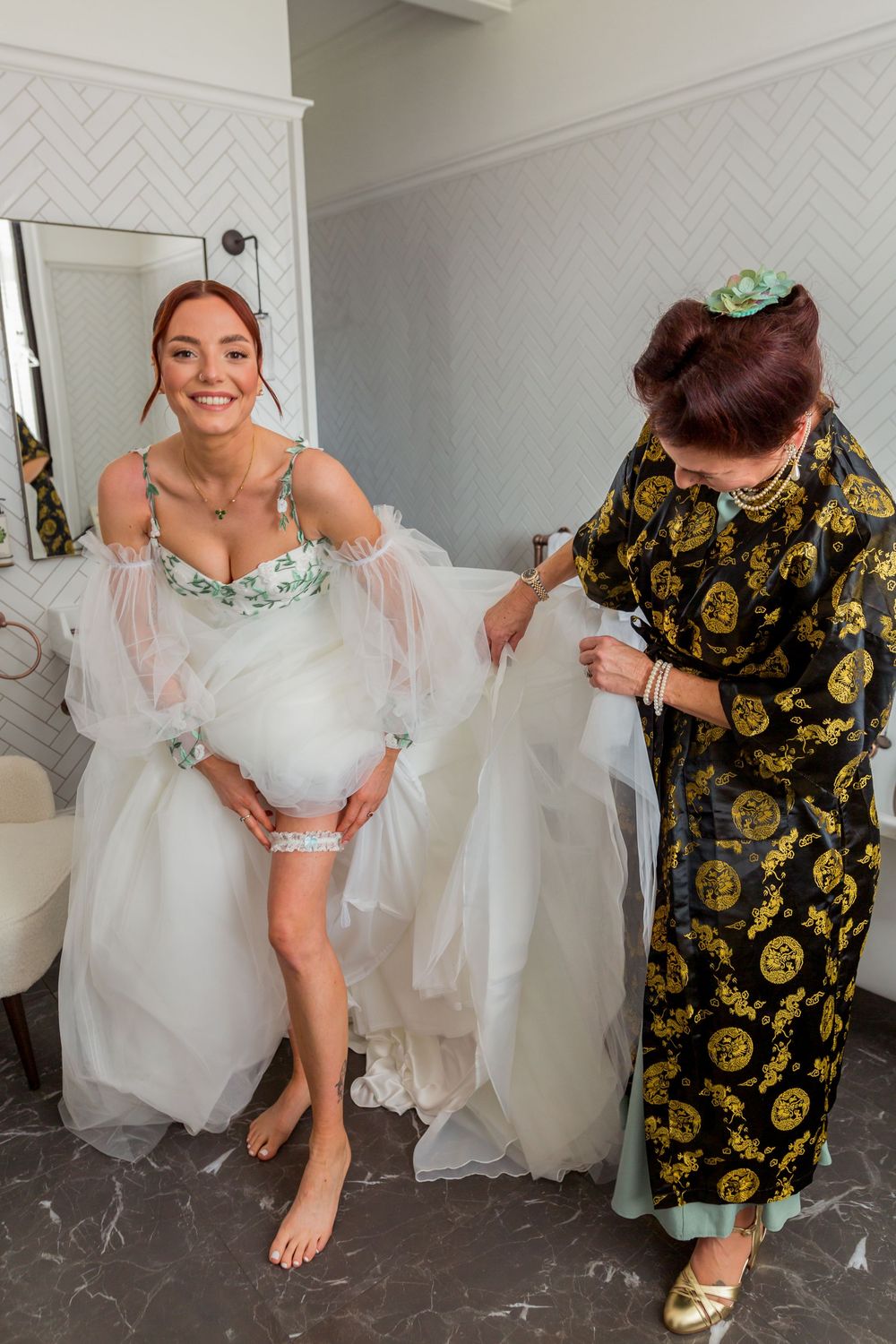 A bride in a white tulle gown getting ready with assistance in a dressing room with patterned wallpaper.