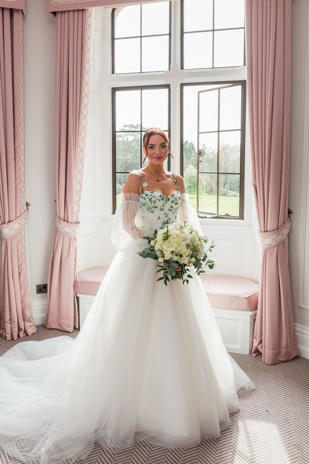 A bride in a flowing white wedding gown stands by bright windows with pink curtains in an elegant room.