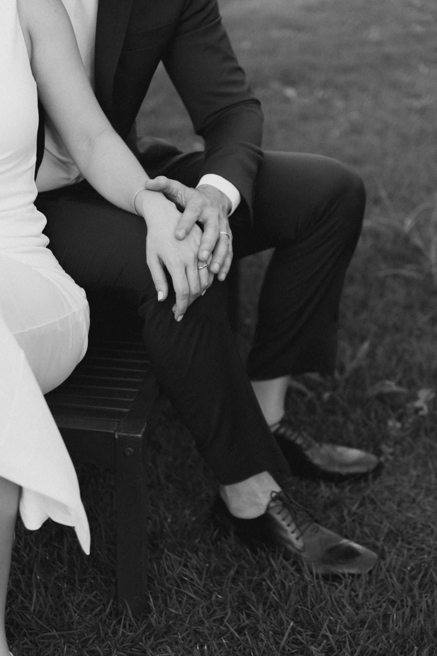 Close-up of clasped hands of a couple in wedding attire sitting on a bench.