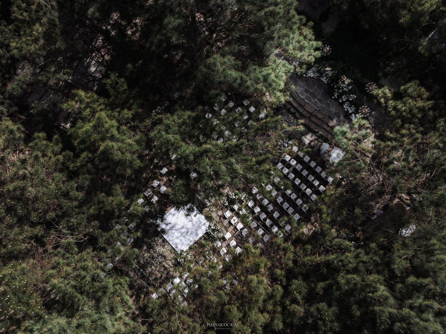 Aerial drone view of outdoor wedding ceremony setup with white chairs nestled among dense forest trees.