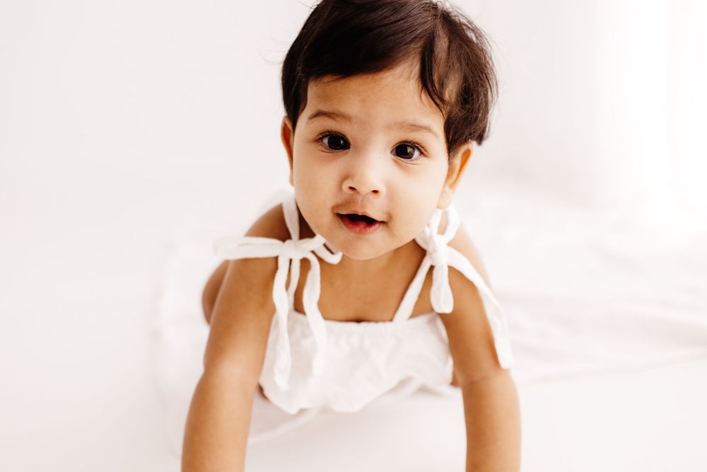Baby in white ruffled dress smiles against minimalist white background.