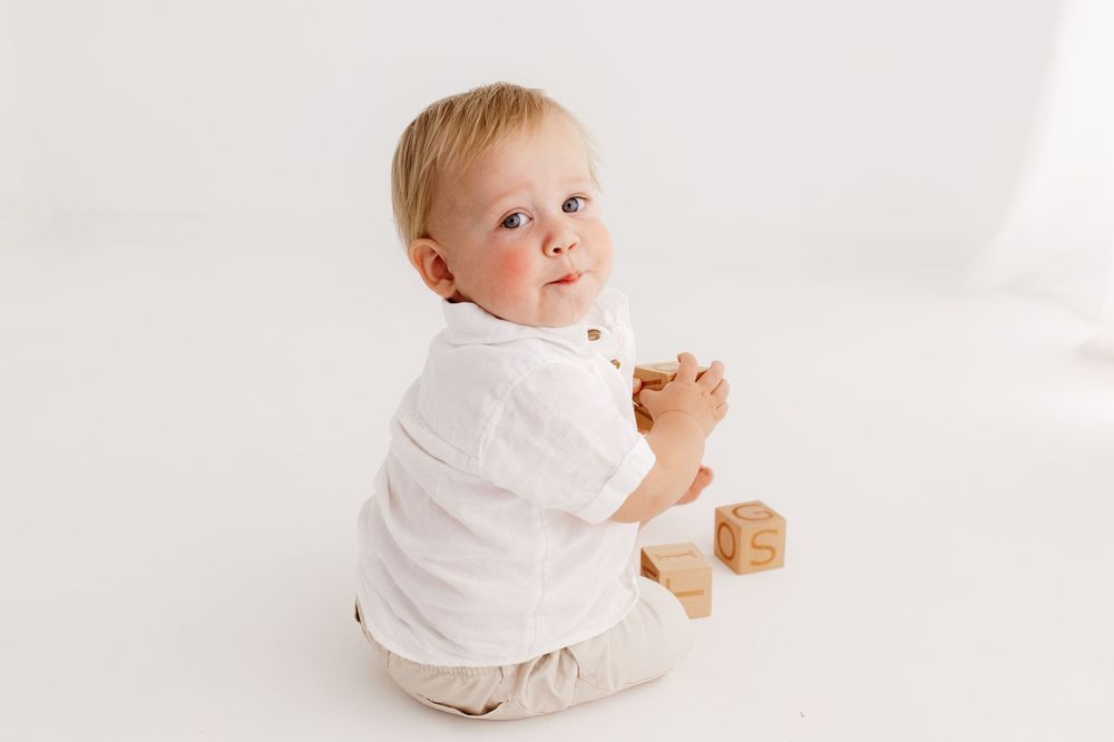 Baby in white outfit plays with wooden block against bright background.