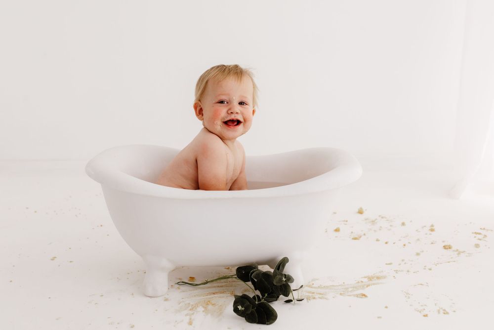 Baby sitting in white bathtub during cake smash cleanup photo session.