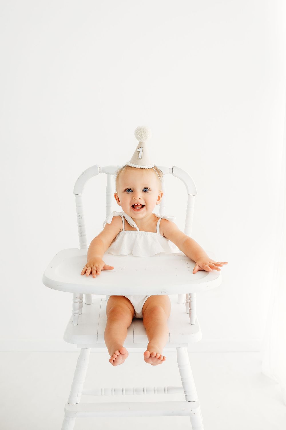 Baby in white dress and party hat sits in antique high chair.