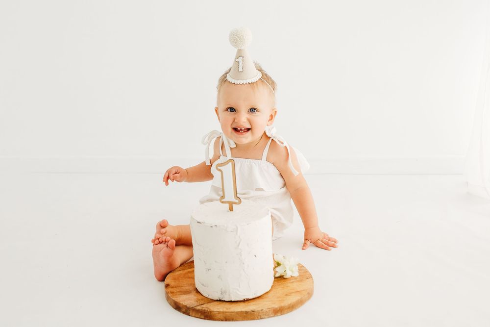 Baby in white dress with party hat sits next to round white cake on wooden board.