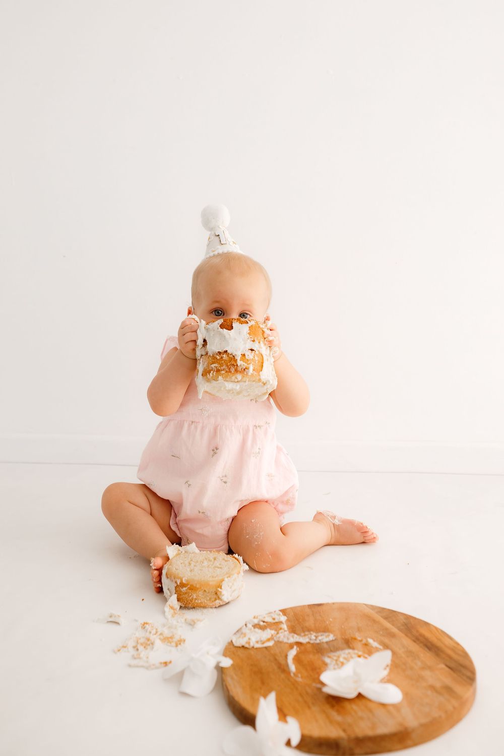 Baby in pink romper playing with cake on wooden board during photoshoot.