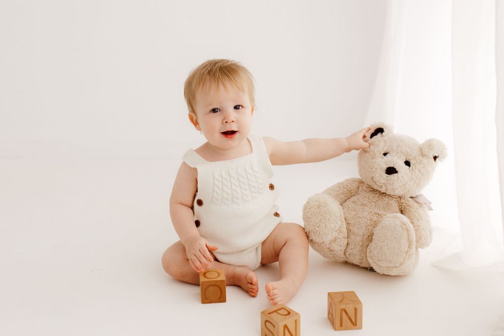 Baby in white onesie reaches for teddy bear during minimalist photo session.
