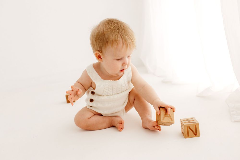 Baby in cream-colored outfit plays with wooden blocks on white floor.