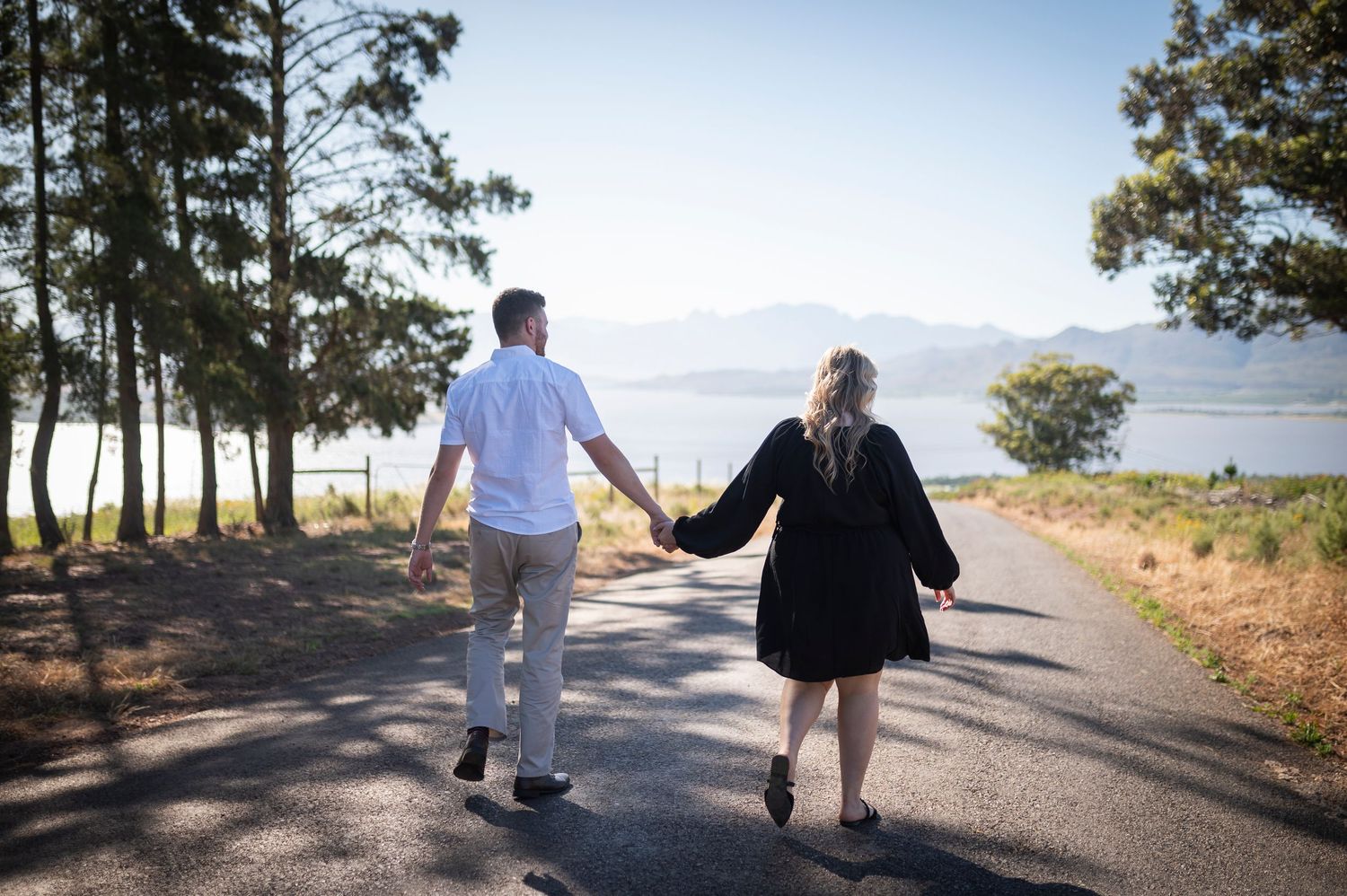 Couple walking hand in hand on a scenic coastal road lined with pine trees overlooking the ocean on a sunny day.