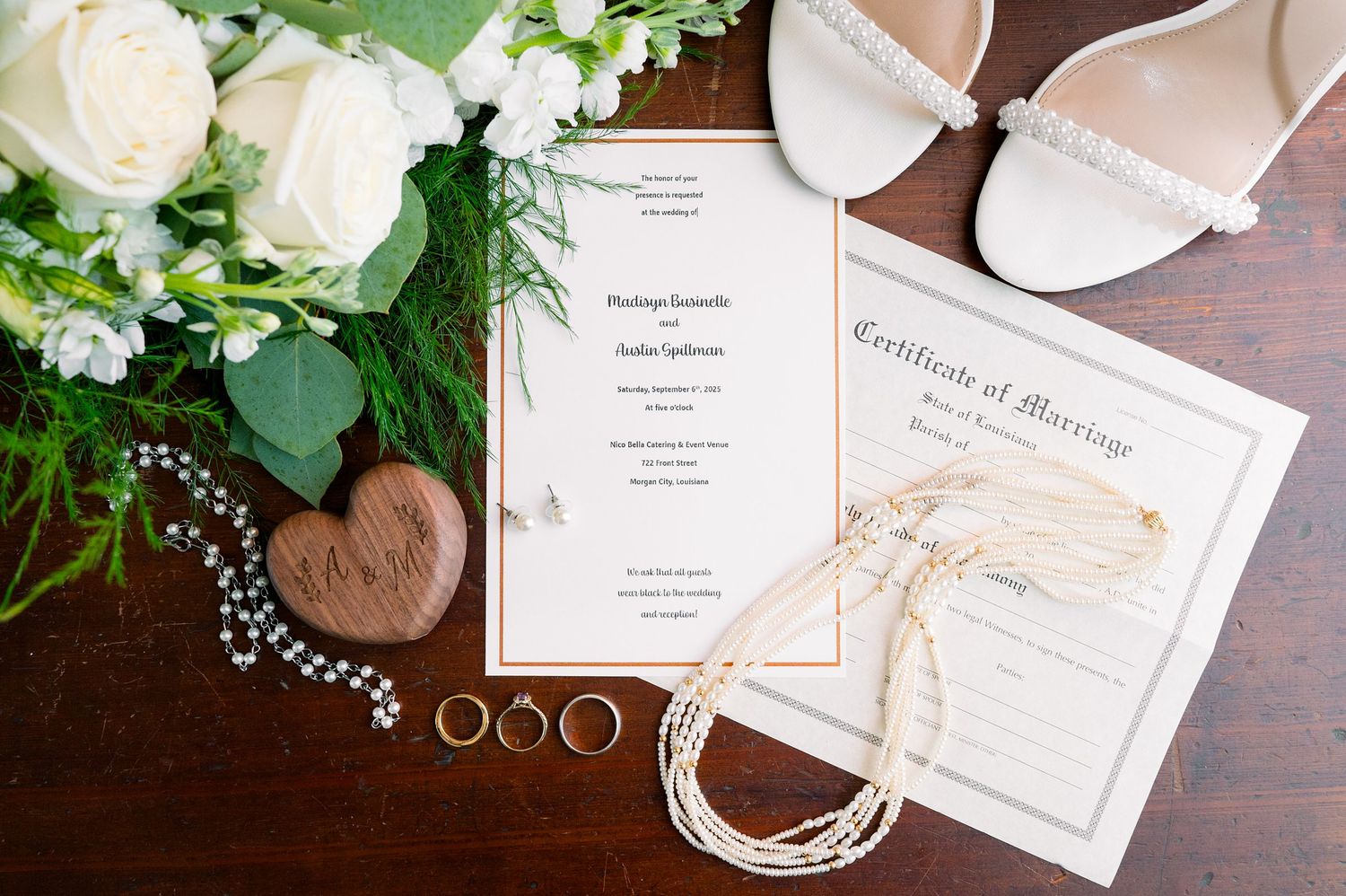 Wedding invitation suite with white roses and gold rings on dark wooden surface.