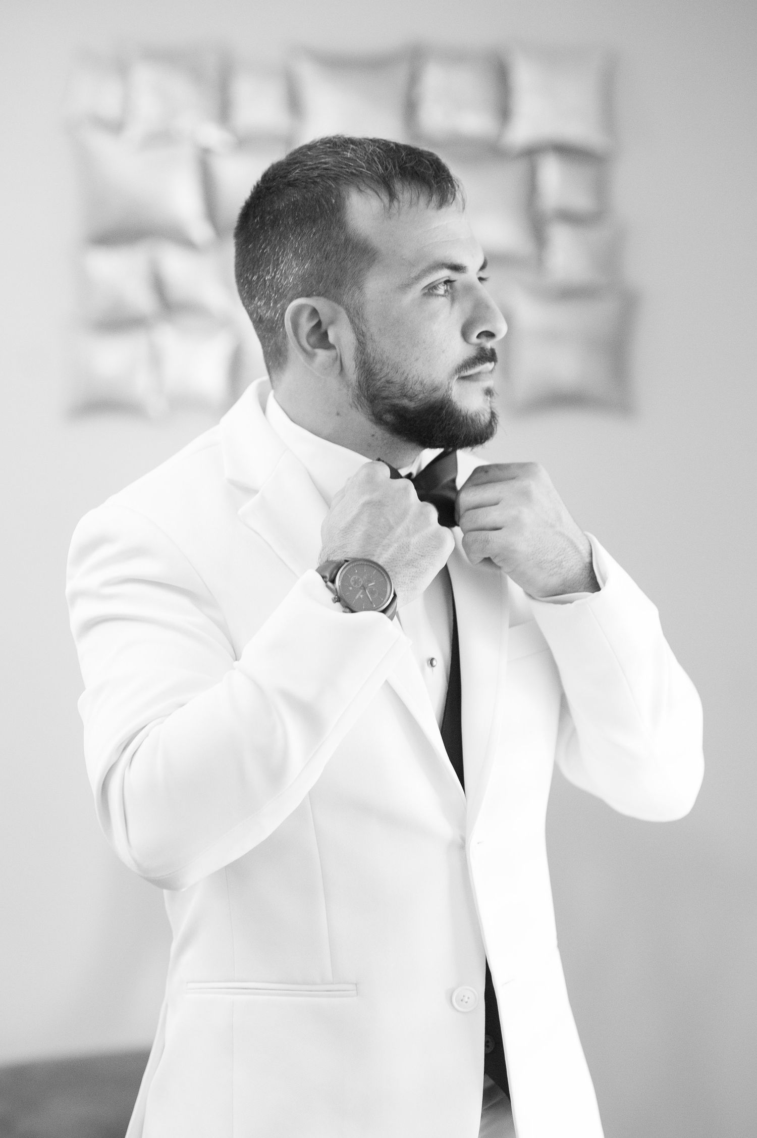 Groom adjusts his white jacket collar in a bright modern setting.