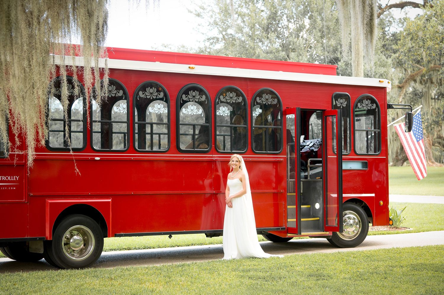 Red vintage trolley bus parked on grass under trees for wedding transportation.
