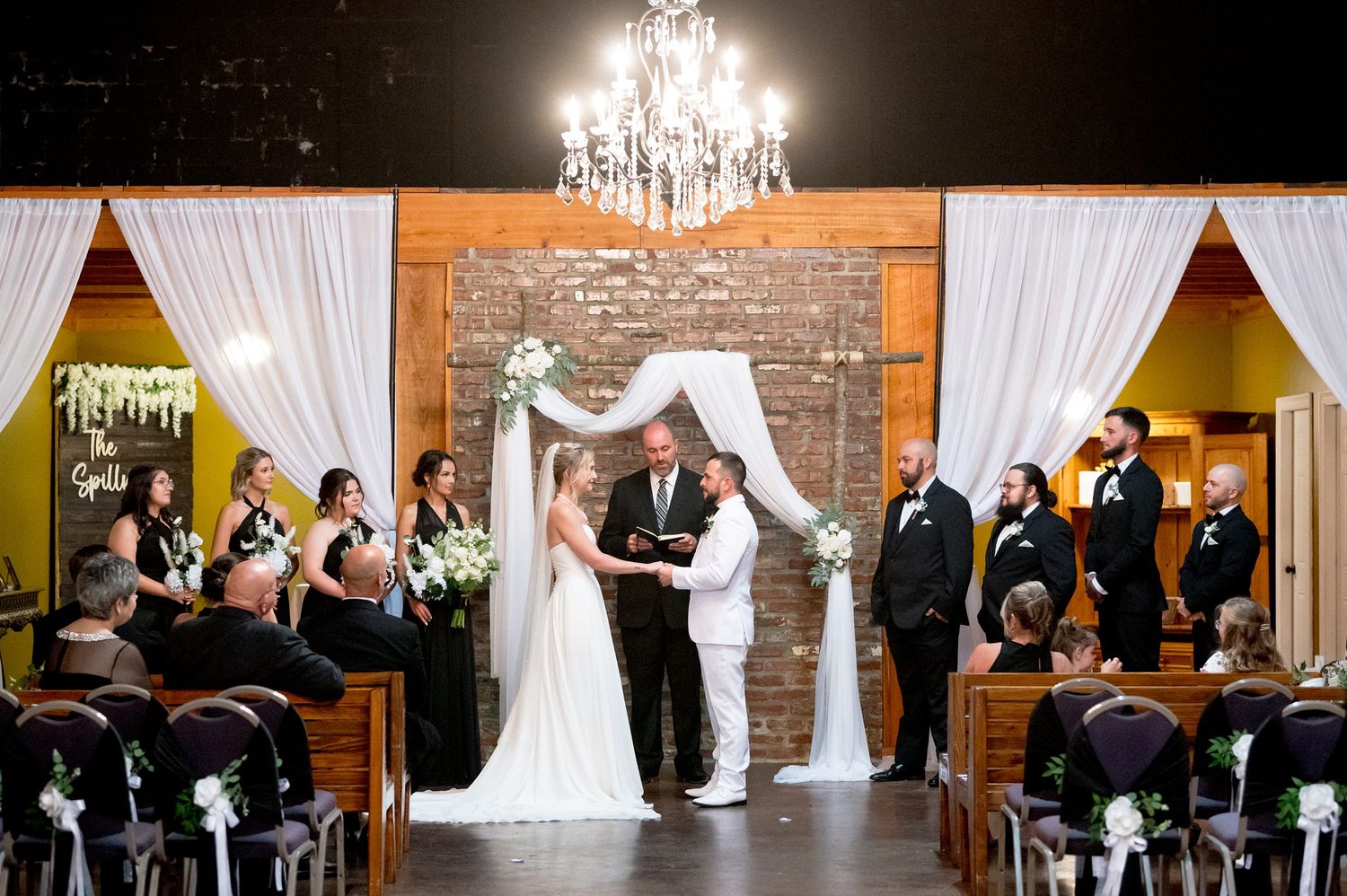 Wedding ceremony space with white draping, chandeliers and exposed brick walls.