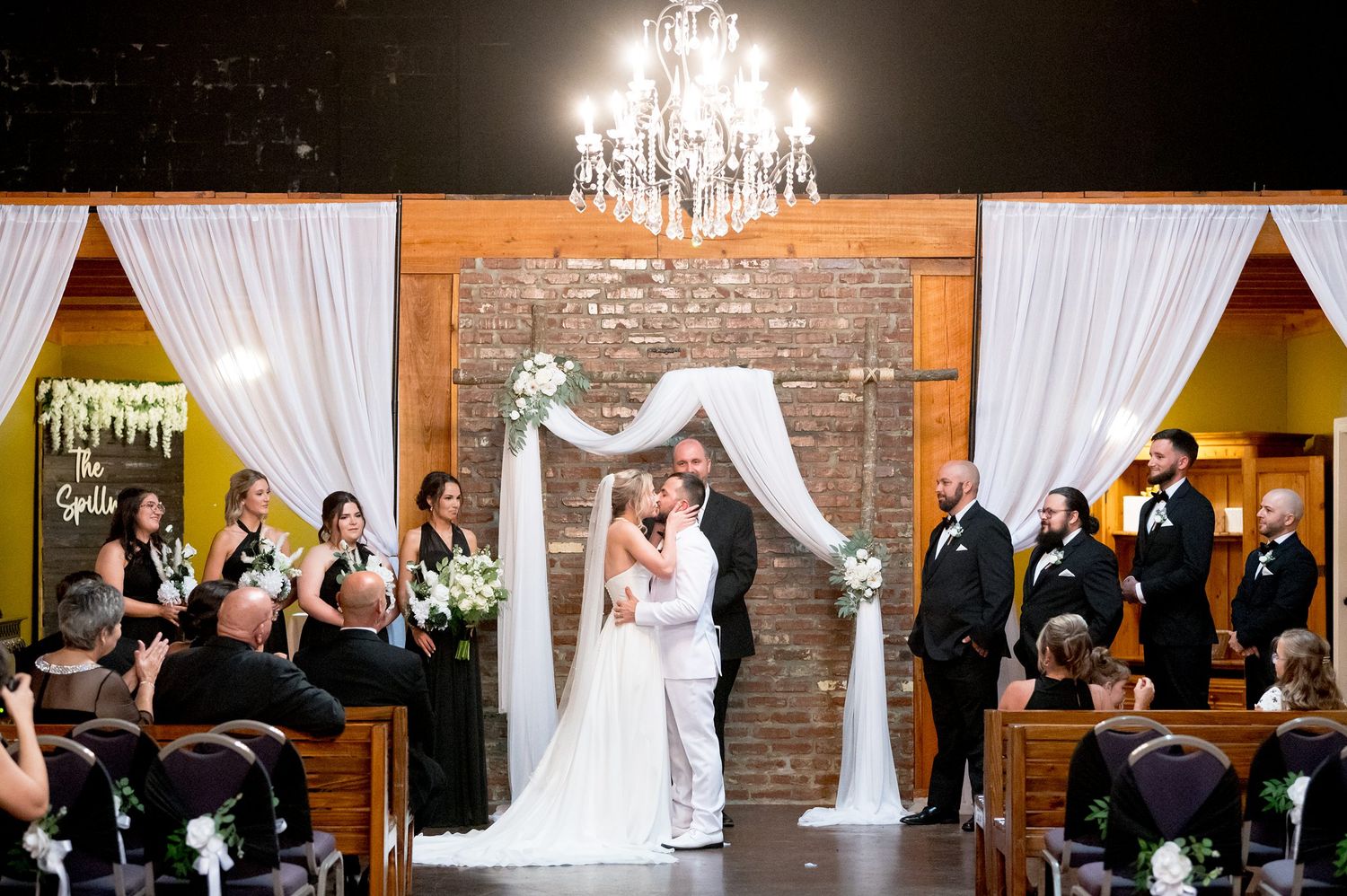 Elegant wedding ceremony under crystal chandelier with white fabric draped arch and brick accent wall.