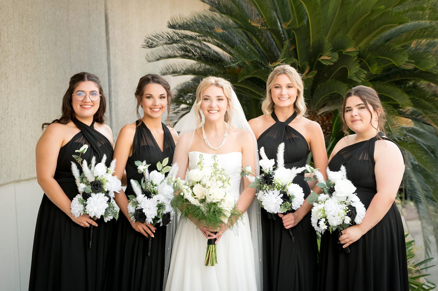 Bridal party in elegant black dresses holding white bouquets posed by palm tree.