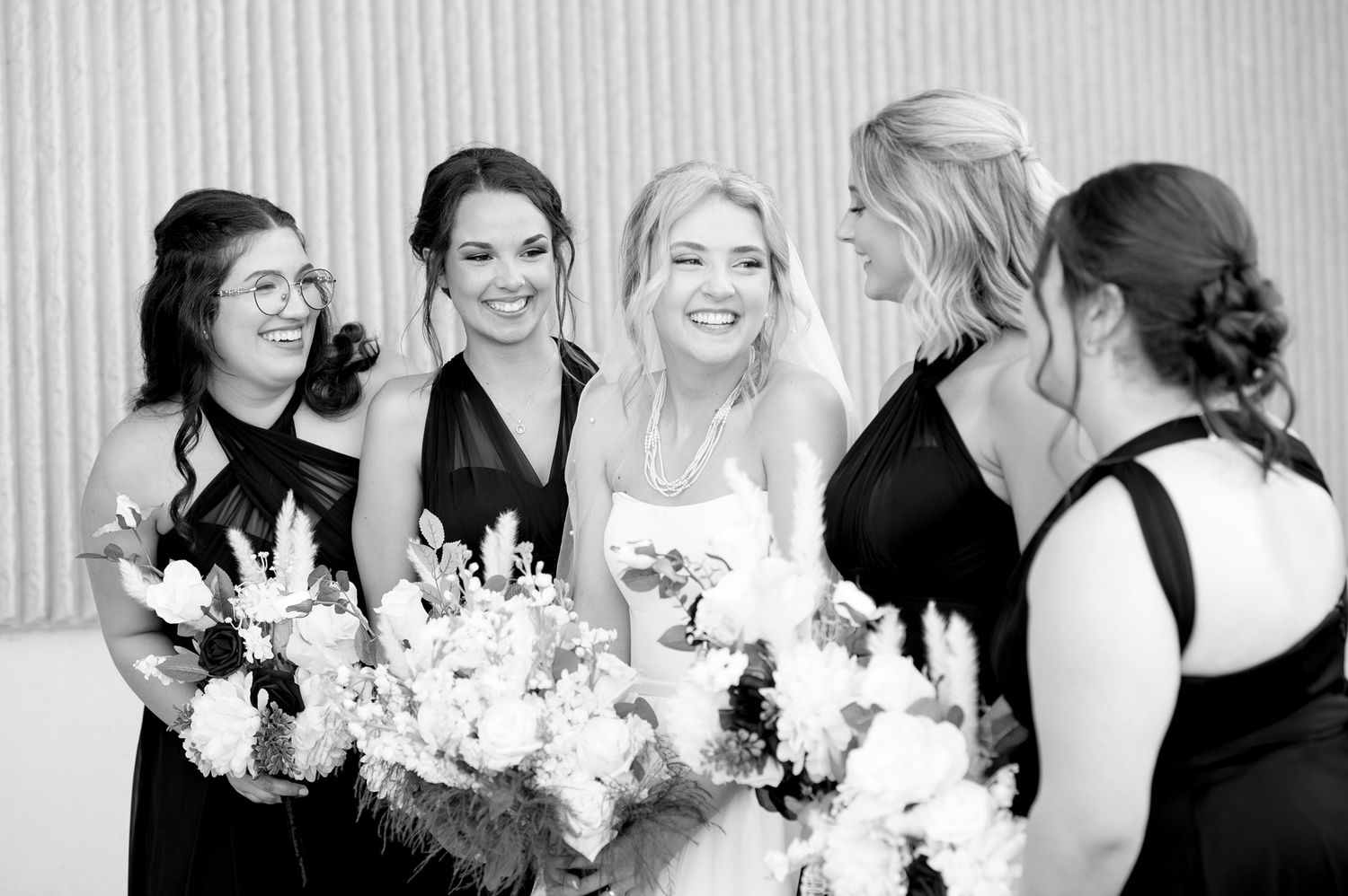 Bridal party in black dresses sharing joyful moment with white floral bouquets.