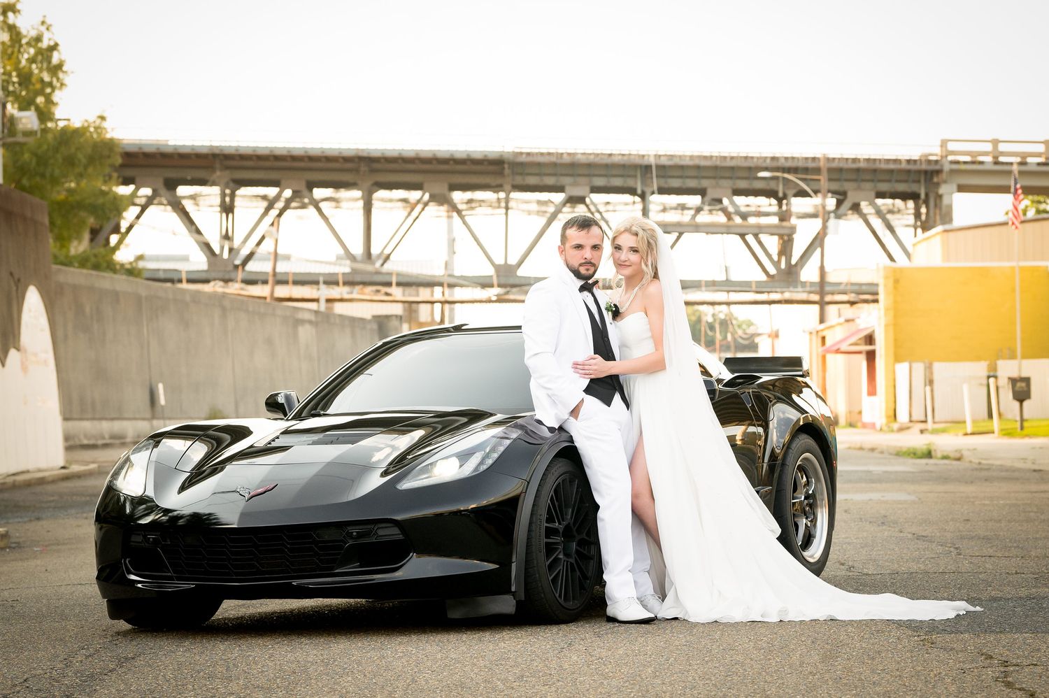 Newlyweds pose with sleek black sports car in urban setting.