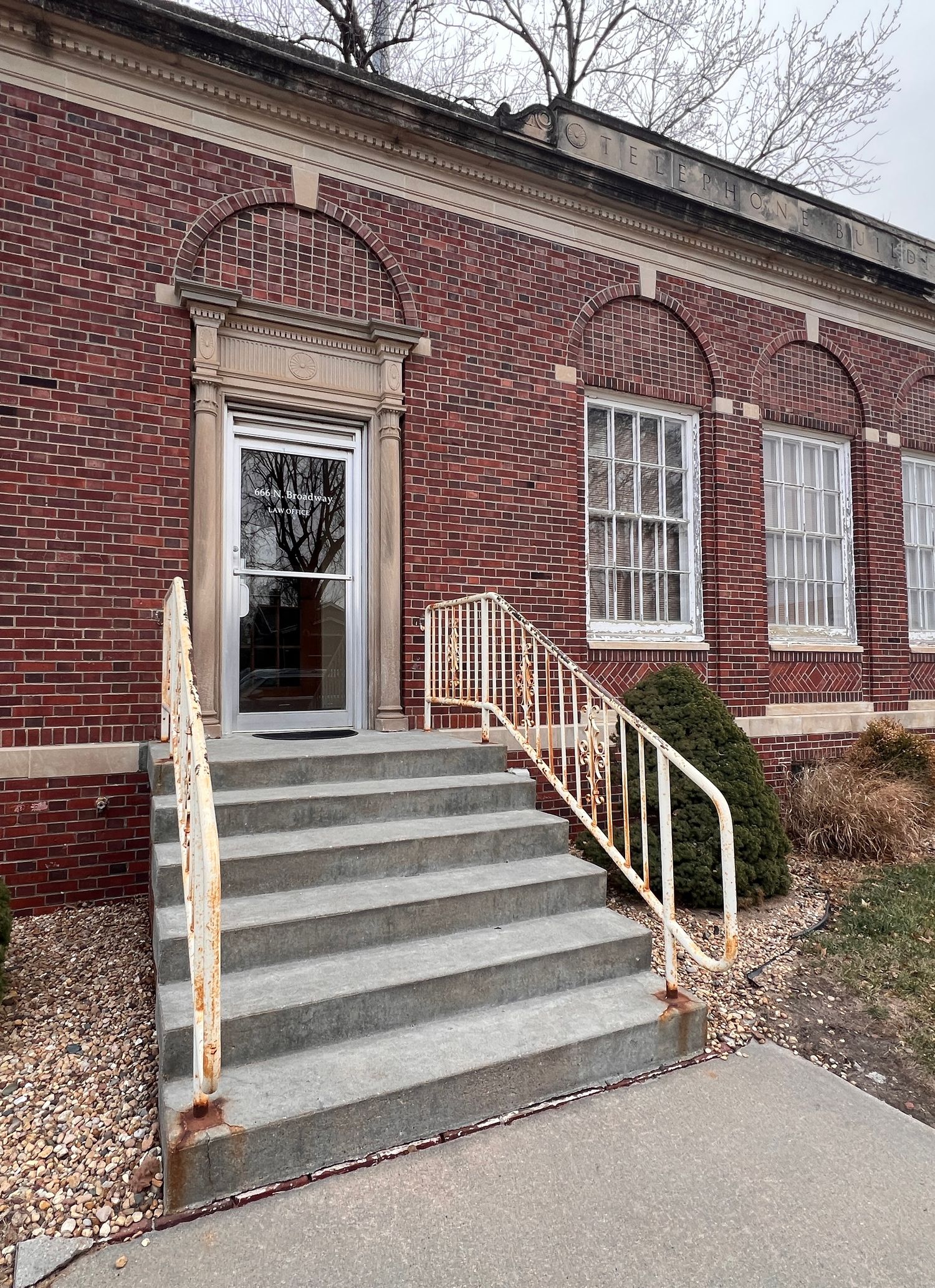 Red brick building entrance with concrete steps and wooden handrails leading to multiple windows.