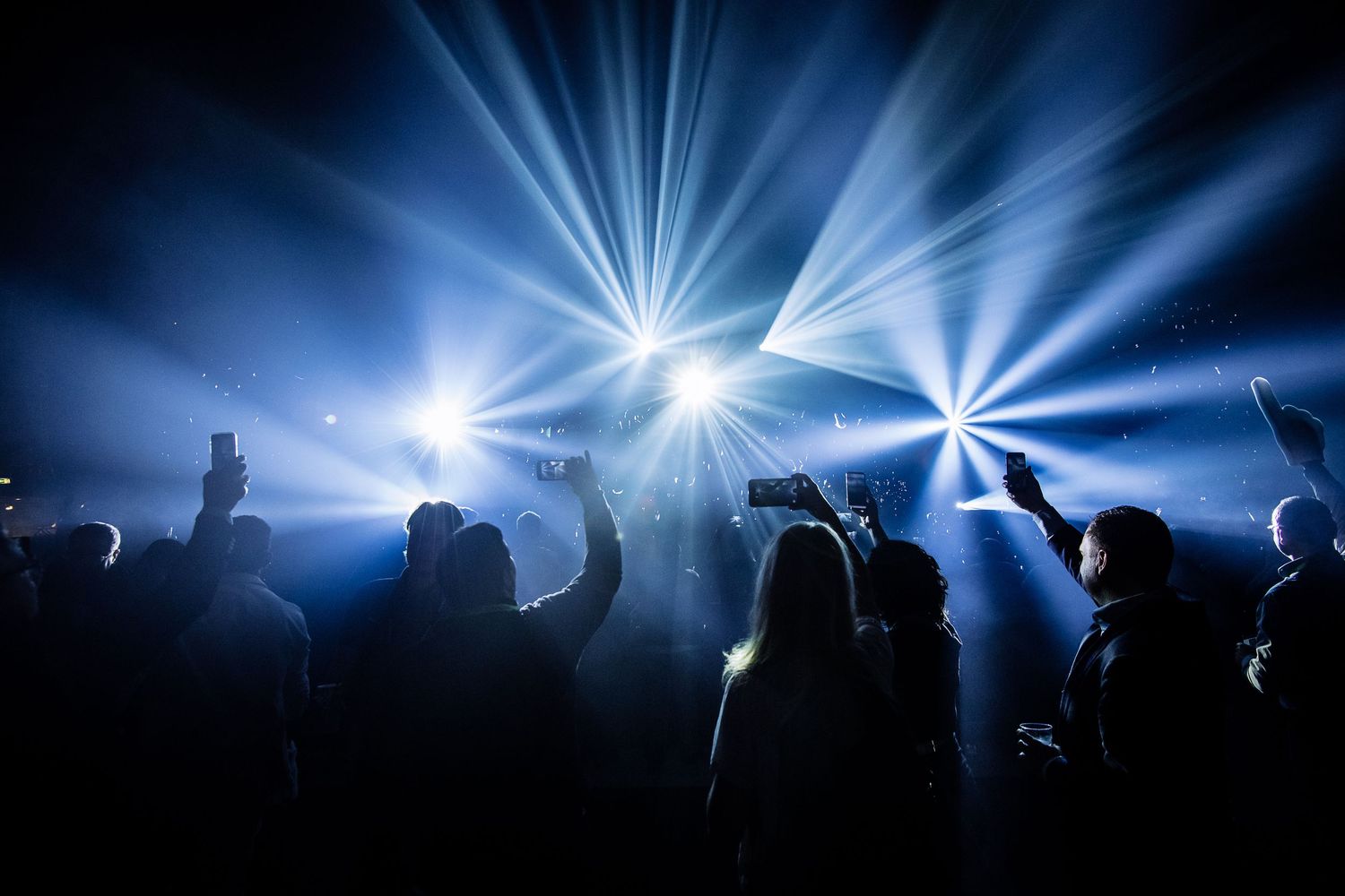 Concert audience silhouettes against bright white stage lighting beams.